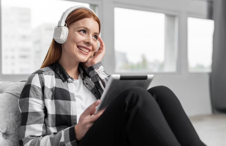 A woman sitting on a sofa, holding a tablet and wearing a headset